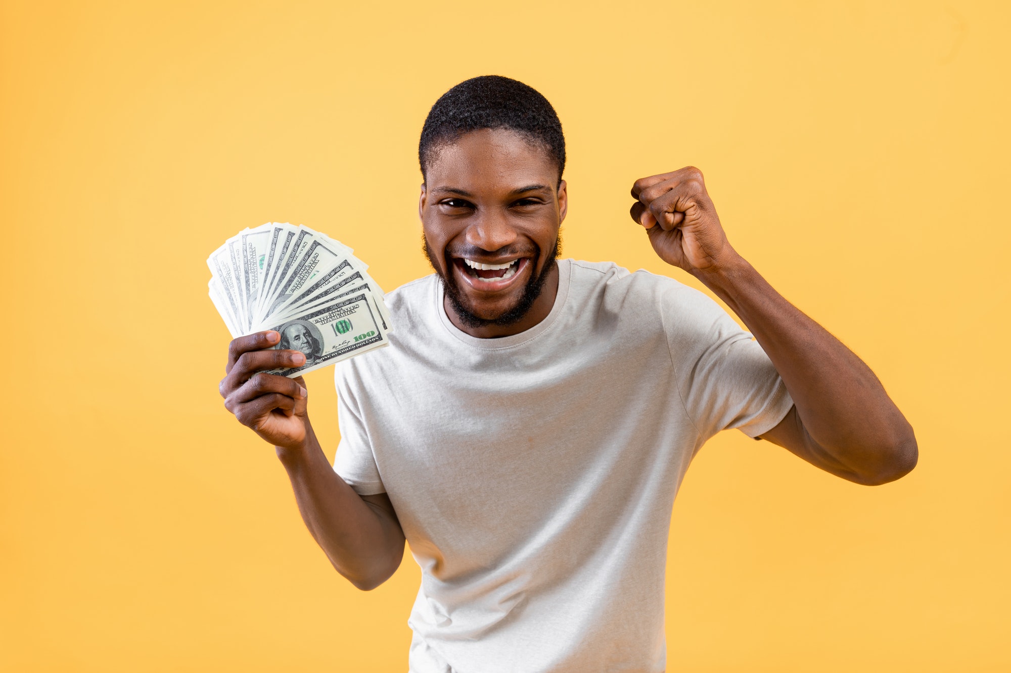 Overjoyed african american man holding lots of money, making YES gesture on yellow studio background
